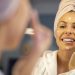 Portrait of beautiful Caucasian woman in towel looking herself in the mirror and applying face cream. She is going through morning beauty routine in bathroom. Skin Care and Hygiene