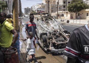 People walk past a burned out car in Dakar, on June 5, 2023, as protest calmed down four days after a court in Senegal sentenced opposition leader Ousmane Sonko, a candidate in the 2024 presidential election, to two years in prison on charges of “corrupting youth” but acquitted him of rape and issuing death threats. (Photo by JOHN WESSELS / AFP)