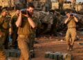 Israeli soldiers carry heavy shells past battle tanks deployed at a position along the border with the Gaza Strip and southern Israel on December 31, 2023, amid the ongoing conflict between Israel and the militant group Hamas. (Photo by Menahem KAHANA / AFP)